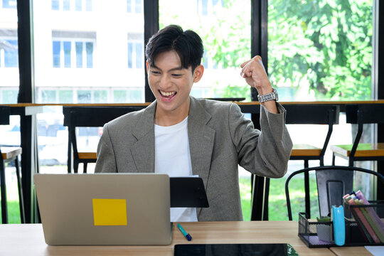 Excited Young Asian Businessman Celebrating Success on Laptop at Modern Office Desk