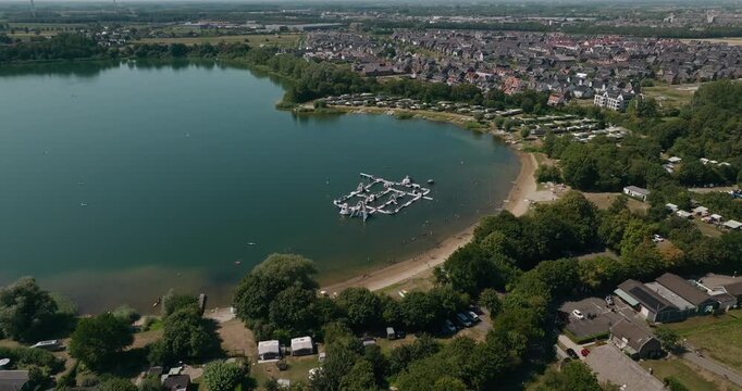 Aerial view of a recreational lake Everstein with a water obstacle course and sandy beach, surrounded by trees and modern Dutch residential area under a clear blue summer sky, Hagestein Hoef en Haag.