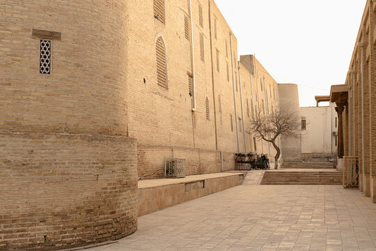 Empty courtyard beside historic brick buildings near Poi Kalyan in Bukhara. Ancient Islamic architecture with lattice windows and stone pavement creates a quiet scene ideal for travel and heritage.