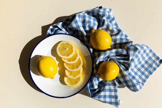 Tranches de citrons jaunes et un citron entier pos&eacute;s sur une assiette &eacute;maill&eacute;e blanche avec un liser&eacute; bleu, un torchon &agrave; carreaux bleus pos&eacute; &agrave; c&ocirc;t&eacute; avec deux citrons, vue du dessus, lumi&egrave;re &eacute;clatante