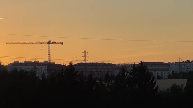 tower crane stands above apartments and transmission pylon at sunset, with hanging hook, warm orange sky, distant skyline, calm urban atmosphere and haze