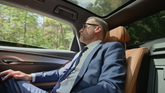 Middle aged Caucasian businessman in suit sitting in backseat of moving luxury car, looking out window. Footage represents executive travel, wealth management, and corporate success concepts