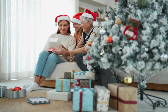 Senior parents opening Christmas present with adult daughter beside decorated tree and wrapped gifts during family holiday celebration at home.
