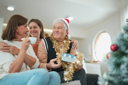 Adult daughter embraces senior parents as father holds Christmas gift, sharing love and happiness together during family holiday celebration at home.