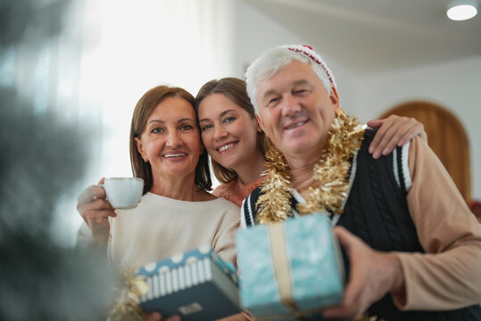 Senior parents and adult daughter smiling together while holding Christmas gifts, sharing affection and joyful family moments during holiday celebration at home.