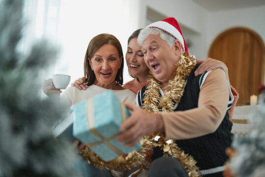 Senior father opens Christmas present with surprised expression while wife and adult daughter watch, sharing excitement and happiness during family gift exchange.