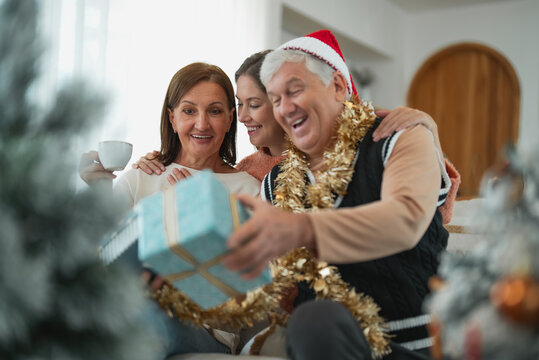 Senior father opens Christmas gift while wife and adult daughter smile beside him, enjoying surprise, gift giving and family togetherness during holiday celebration.