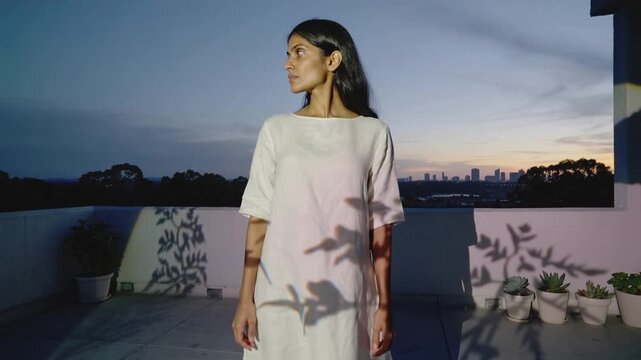Standing Indian model turning head left in white midi dress roof moving plants casting leaf shadows