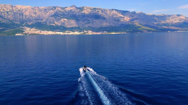 A lone motorboat glides across calm adriatic waters, set against majestic mountains. In the summer sun, a couple leisurely explores the stunning coastline near a small beach town.