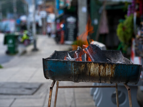 Burning charcoal brazier on Asian sidewalk with blurred busy street behind, street food preparation