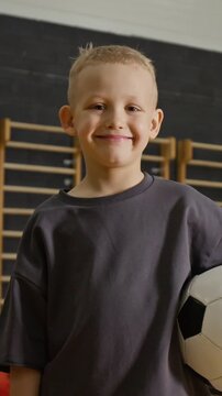 Portrait of smiling Caucasian boy holding soccer ball in school gym. Footage is perfect for youth sports marketing or physical education advertising campaigns, vertical
