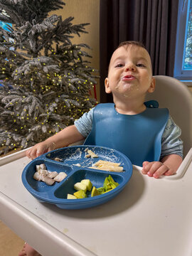 Messy toddler eating in high chair by christmas tree.