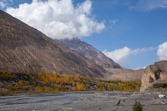 View of Sost village featuring golden autumn trees along the riverbank, rugged mountain slopes, and a clear blue sky with white clouds in Gilgit-Baltistan, Pakistan.