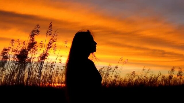 Silhouette of a woman with long hair standing outdoors in profile against a vibrant orange and yellow sunset sky with tall reeds.