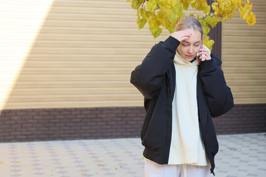 Young Woman Stands Outside Beside Yellow Autumn Leaves And Sunlit Wall, Holding Mobile Phone To Ear With Hand On Forehead And Eyes Closed, Expressing Stress, Concern, And Private