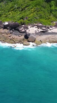 Vertical aerial pull back view revealing the rocky shore of Grande Anse on Mahe Island in Seychelles with turquoise water and tropical coastline.