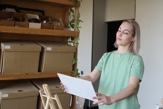 Blonde Woman In Mint Shirt Stands Beside Wooden Shelves Filled With Cardboard Boxes, Inspecting Blank Sheet In Bright Indoor Workspace With Natural Light, Houseplant, And Tidy Creative