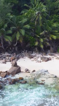 Vertical aerial pull back view revealing Petite Police Beach in Seychelles with turquoise water, white sand, granite rocks and lush tropical vegetation.