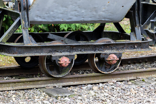 Narrow gauge wagon wheelset on rail track with rusted axle and steel wheels visible. Close view shows gravel ballast and corroded metal for industrial parts identification and restoration reference.
