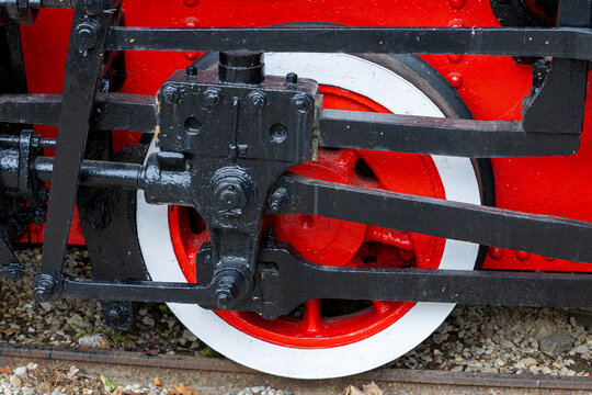 Steam locomotive driving wheel with connecting rod crankpin valve gear and piston linkage on narrow gauge track with ballast. Close crop shows mechanical detail for restoration maintenance.
