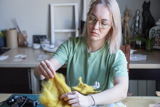 Adult Woman With Glasses Carefully Shaping Yellow Wool Into Felt Figurine At Worktable In Home Studio, Focused Handmade Textile Craft Scene With Soft Natural Light, Creative Hobby