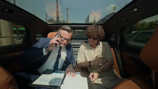 Middle aged Caucasian businessman talking on smartphone and reviewing documents with young adult colleague in backseat of moving car. Perfect for corporate travel and mobile office concepts