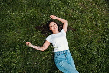 Fototapeta premium Young woman relaxing on green grass, smiling and enjoying a sunny outdoor moment with casual jeans and white t-shirt, showing a floral arm tattoo and carefree summer vibe