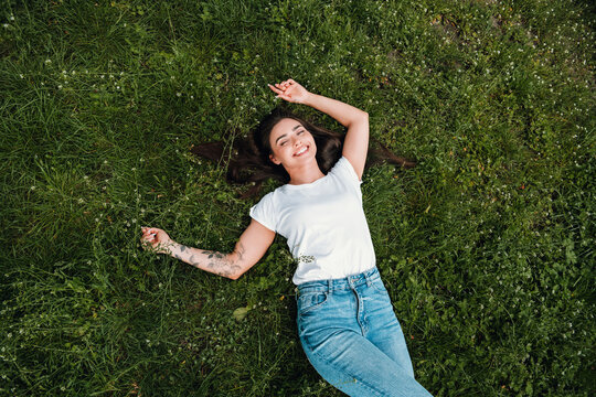 Young woman relaxing on green grass, smiling and enjoying a sunny outdoor moment with casual jeans and white t-shirt, showing a floral arm tattoo and carefree summer vibe