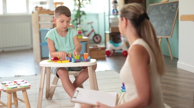 Attentive male kid assembling colorful train blocks near specialist taking notes in preschool room