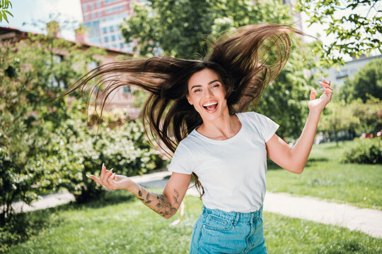 Joyful young woman flipping her long hair and smiling in a sunny city park wearing a white t-shirt and denim skirt with visible tattoo
