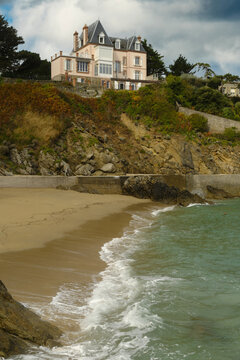 A grand coastal villa overlooks a sandy beach and turquoise sea in Dinard, Brittany, under a dramatic cloudy sky.