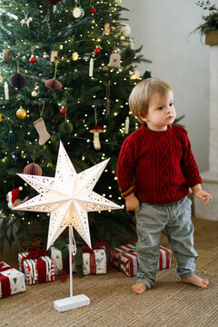 Cute Toddler Boy in Red Knit Sweater Standing Barefoot Near Decorated Christmas Tree. Concept of Holiday Warmth and Winter Magic