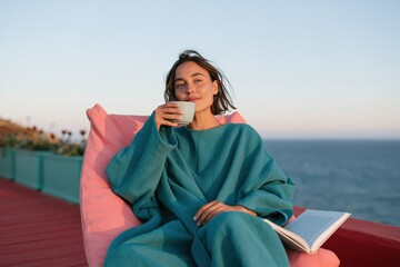 Fototapeta premium Woman relaxed on a pink lounge chair drinking coffee and reading by the ocean at sunset for calm seaside background and lifestyle design