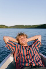 Fototapeta premium Smiling young man reclining on a boat bow with arms behind head at a lake in striped shirt for summer portrait background and travel poster