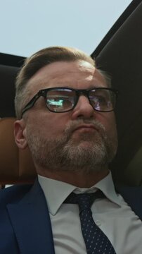 Vertical shot of middle aged Caucasian businessman in suit sitting in car, answering smartphone and talking. Footage is ideal for corporate communication and executive lifestyle marketing