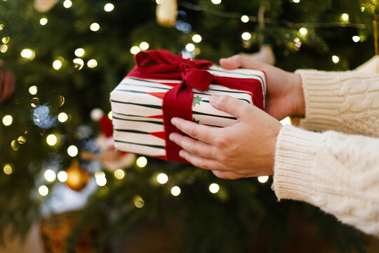 Hands in Knit Sweater Holding Wrapped Christmas Gift Box with Red Ribbon on background of golden xmas tree lights bokeh. Christmas Present Exchanging. Merry Christmas