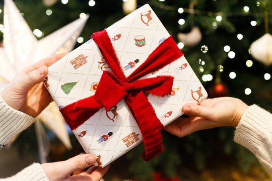 Close up of hands passing wrapped Christmas gift with red ribbon near glowing tree lights. Holiday gift giving tradition, family care, and festive winter celebration. Merry Christmas