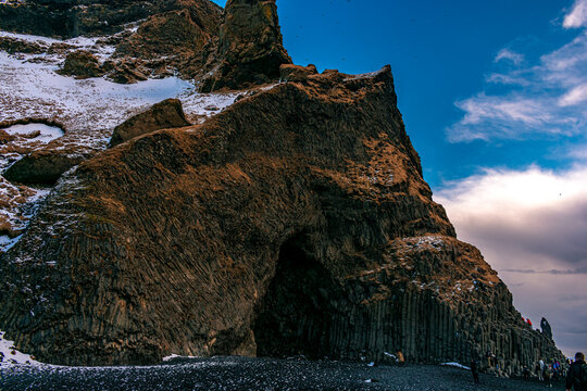 View of the dramatic basalt columns at Reynisfjara beach with snow-dusted volcanic cliffs and black sand under a bright blue sky, Iceland.