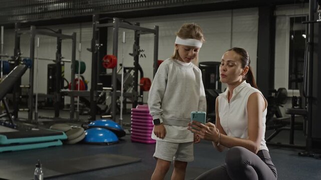 Caucasian woman and Caucasian child looking at smartphone in gym, tracking workout progress and flexing muscles. Perfect for promoting family fitness apps or healthy lifestyle campaigns