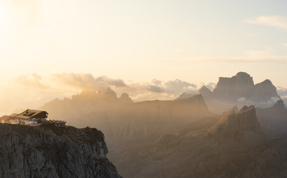 View of a mountain hut perched on a rocky cliff edge with the dramatic peaks of the Dolomites glowing in the warm light of sunset in Veneto, Italy.