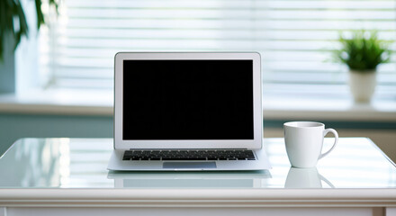Fototapeta premium Modern silver laptop and white coffee cup on glass desk in bright home office with window blinds and plant, minimalist workspace with calm atmosphere