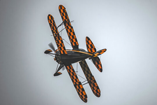 View of an aerobatic biplane with a distinctive black and orange checkerboard pattern performing a maneuver against a cloudy sky in Cascais, Lisbon, Portugal.
