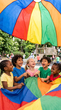Children sharing a large colorful parachute game in an outdoor playground, cooperation, movement and laughter, diverse representation, simple park background, Children's Day