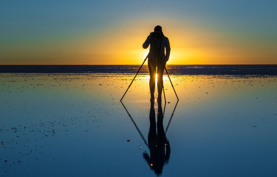 View of a photographer silhouetted against the vibrant sunset sky while standing with a tripod on the reflective surface of the Uyuni Salt Flat in Uyuni, Potosi Department, Bolivia.