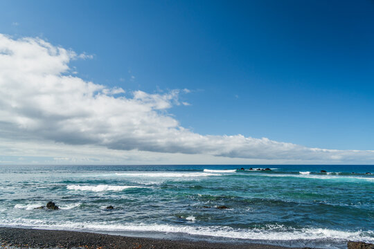 View of the coastal landscape of Las Galletas with dark volcanic sand, turquoise ocean waves, and a clear blue sky with a long cloud formation in Las Galletas, Canary Islands, Spain.