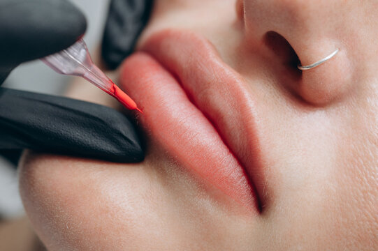 Macro close up of a permanent makeup artist performing a lip blush tattoo procedure with a PMU machine in a professional beauty salon.