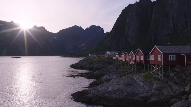 Sun shines brightly over distant mountains and water while wooden rorbu fisherman cabins stand on stilts along the rocky shoreline in Lofoten, Norway.