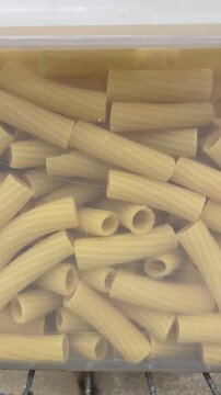 Vertical closeup of uncooked tube shaped pasta stored in transparent plastic food container. Dry pasta neatly arranged for kitchen storage highlights pantry organization, everyday cooking ingredients.