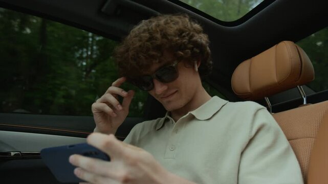 Young adult Caucasian man playing mobile game on smartphone in backseat of moving car. Footage is suitable for mobile app or travel entertainment ads