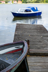 Weathered rowboat moored beside a wooden dock, with a blue motorboat softly blurred on the calm lake in the background. © Do